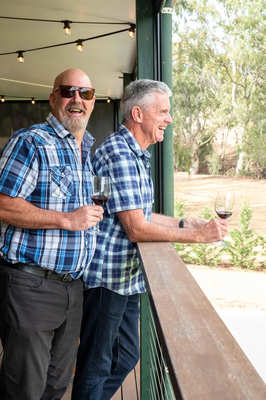 Two guests holding glasses of Barossa red on a cellar-door verandah with vines behind them