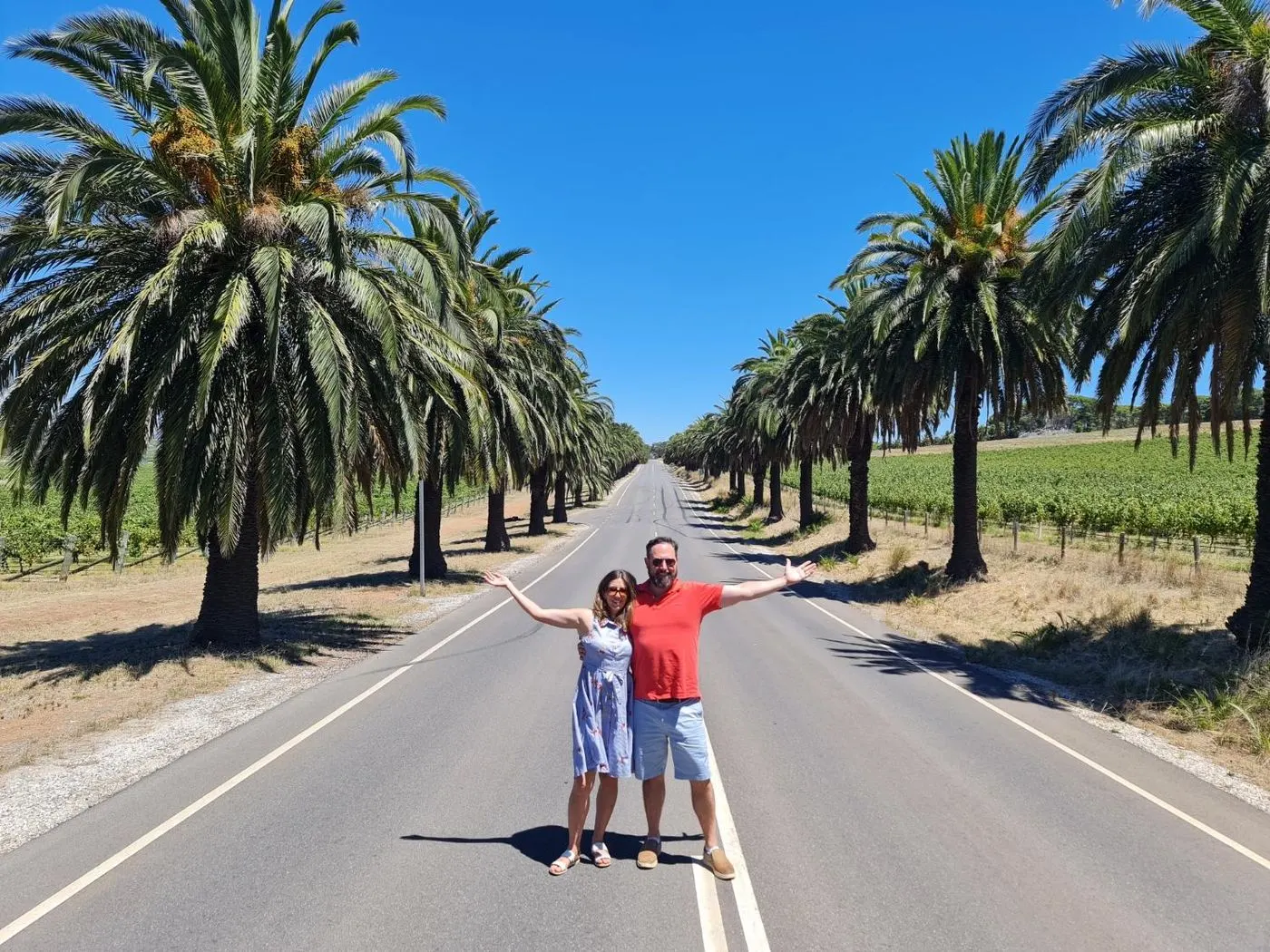 Two guests standing arms wide in a palm-lined road through the Barossa vineyards